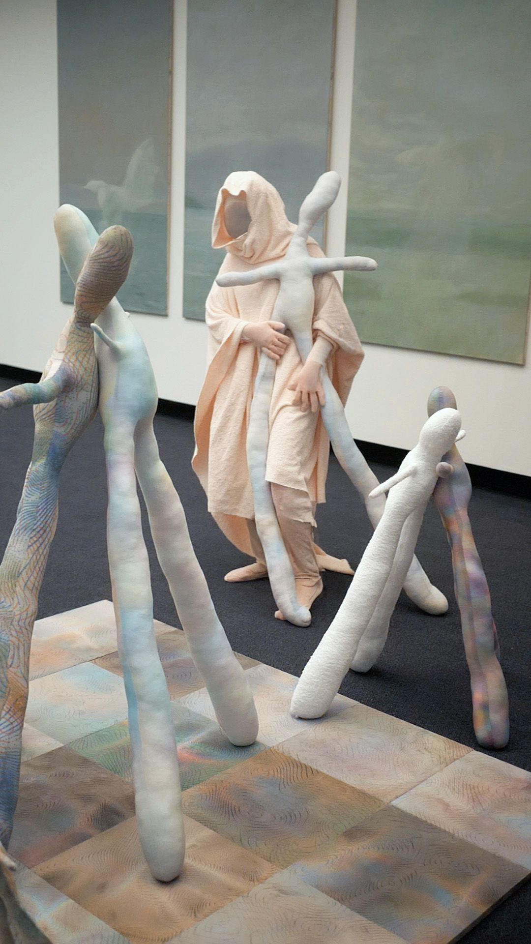 Tejzt's performance 'Variantenraum 934' – A hooded figure dressed in pale fabric interacts with elongated, humanoid sculptures in an abstract spatial arrangement, evoking a ritualistic sorting of potential realities.