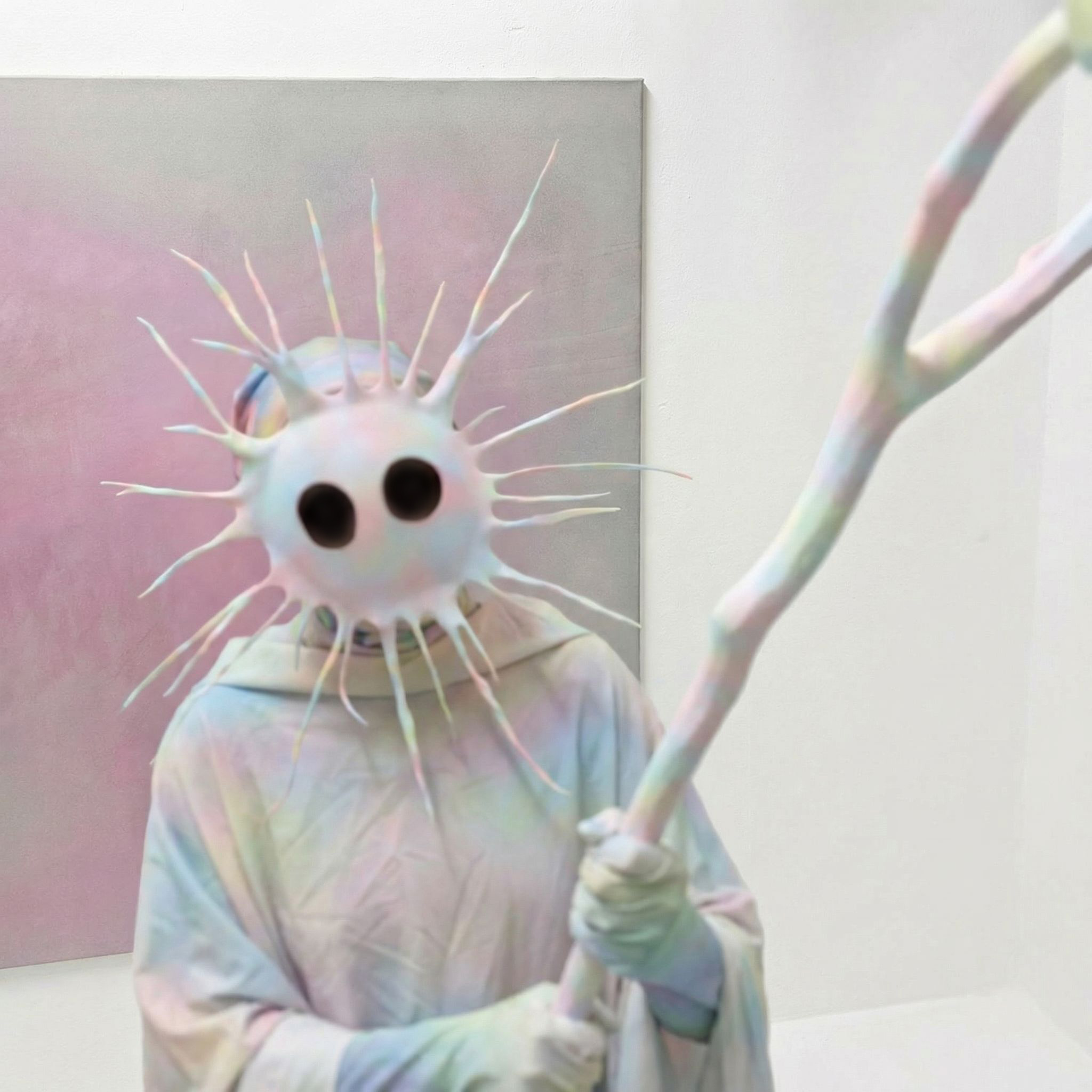 Tejzt – Efee figure in pastel robe with spiked mask and staff, exhibition image (2026)
